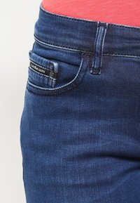 Vaqueros de mezclilla de color azul oscuro con ligeras desgastes, con un bolsillo frontal, etiqueta de marca y corte recto. Parte superior roja visible en el fondo.
