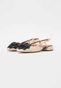 Beige leather flats with a black leather flower embellishment, closed toe, and crisscross ankle straps on a low block heel.