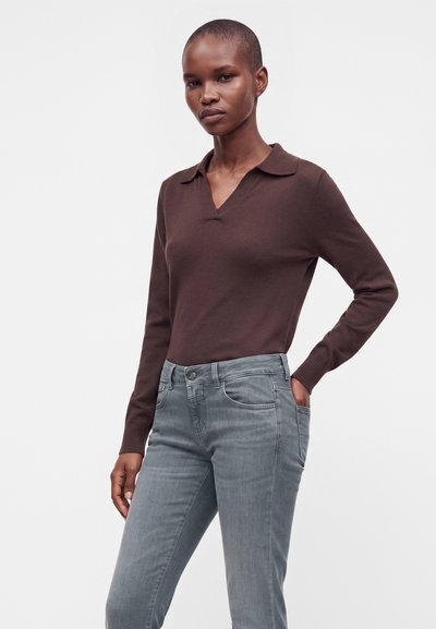Femme aux cheveux courts portant un haut marron à manches longues avec col et un jean bleu clair, debout avec une main dans la poche sur un fond uni.
