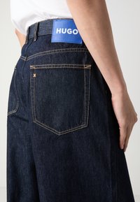Tmavě modré džíny z denimu s vysokým pasem, vybavené modrou nášivkou HUGO na zadní straně, oranžovými švy a dvěma zadními kapsami.