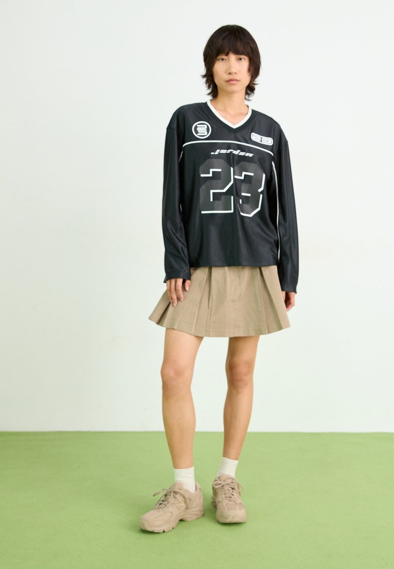 Schwarzes Langarmtrikot mit der Nummer 23, kombiniert mit einem beige gefalteten Rock. Getragen mit beigen Turnschuhen und weißen Socken. Grüner Boden.