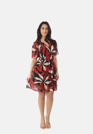 Kobieta stojąca, ubrana w sukienkę do kolan w duże kwiatowe wzory w kolorach czerwonym, czarnym i beżowym, oraz przejrzyste szpilki na białym tle.