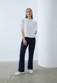 Hvid top med hulmønster og flæseærmer, parret med mørkeblå jeans og hvide sneakers. Minimalistisk design og afslappet pasform.