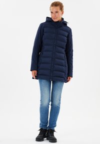 Marinblå vadderad jacka med huva, med en central dragkedja och två sidofickor, som bärs över blåjeans och svarta stövlar. Slät, elegant textur.