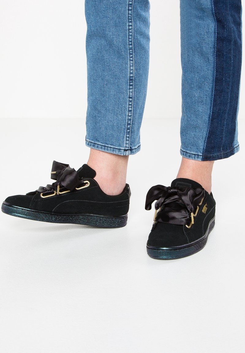 Jambes portant un jean bicolore bleu et des baskets en daim noir avec des lacets en ruban satin noir brillant, debout sur une surface blanche.