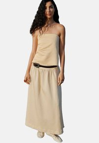 Top beige senza spalline abbinato a una gonna lunga coordinata, con un punto vita arricciato e una texture delicata, completato da una cintura scura.