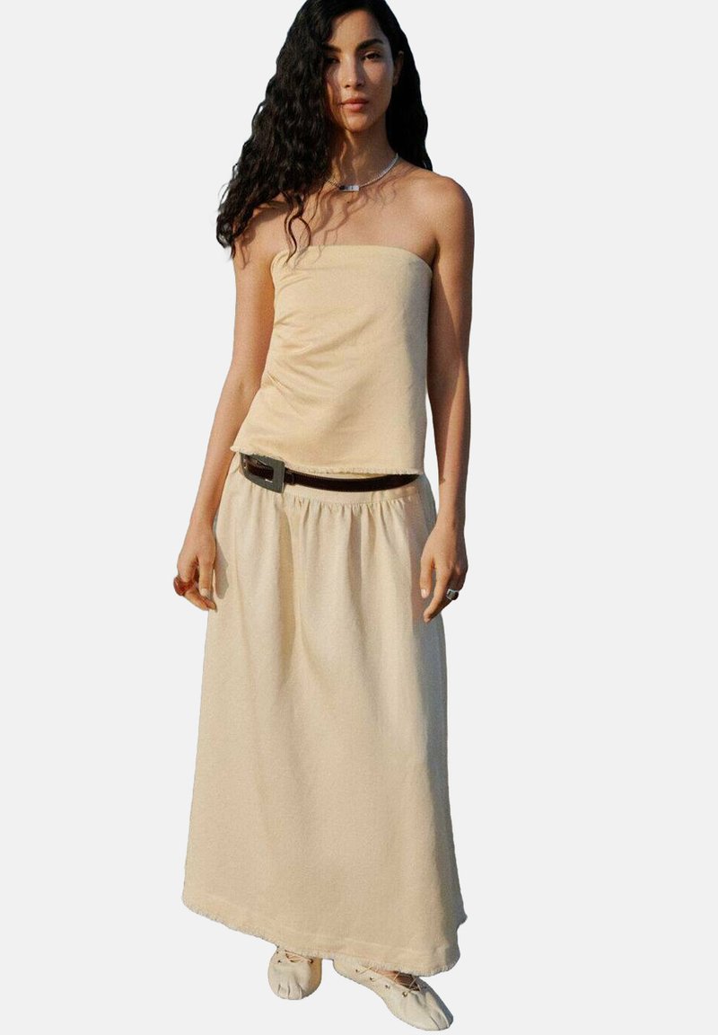 Top beige senza spalline abbinato a una gonna lunga coordinata, con un punto vita arricciato e una texture delicata, completato da una cintura scura.