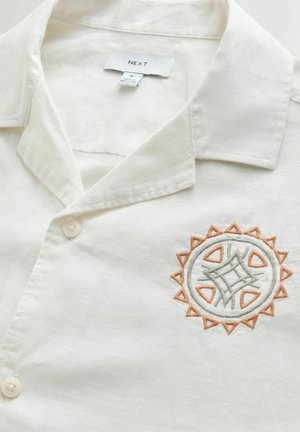 Short-sleeve shirt in light cream fabric, featuring a collar, button closure, and a circular embroidered design in green and orange.