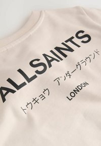 Hellbeige Sweatshirt mit geribtem Halsausschnitt, darauf in auffälligem Schwarz der Schriftzug: "ALLSAINTS" und "LONDON" sowie zusätzliche Zeichen darunter. Weicher, strukturierter Stoff.
