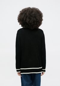 Osoba o kręconych włosach, ubrana w czarny sweter w prążki z białymi paskami na mankietach i dole, w połączeniu z niebieskimi dżinsami, stojąca tyłem na białym tle.