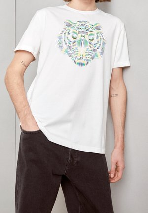 Person trägt ein weißes T-Shirt mit einem mehrfarbigen, stilisierten Tigergesicht, kombiniert mit dunkler Hose, steht mit einer Hand in der Tasche.