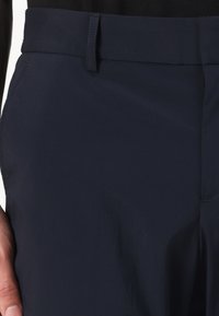 Detail pasu osoby, která má na sobě tmavě modré kalhoty s poutky na pásek a bočními kapsami, ruka spočívá poblíž kyčle.