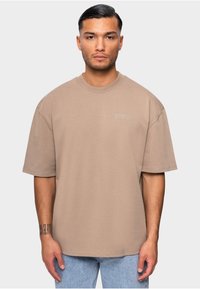 T-shirt beige surdimensionné à manches courtes, avec un col rond et le logo subtil "DROPSIZE" sur la poitrine. Le tissu semble doux et lisse.