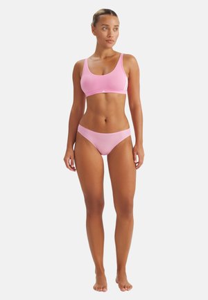 Frau steht barfuß, trägt einen rosa Sport-BH und passende Bikini-Unterwäsche, schaut leicht nach links vor einem weißen Hintergrund.