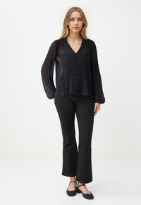 Schwarze plissierte Bluse mit Knopfleiste, V-Ausschnitt, langen Ärmeln und lockerer Passform, kombiniert mit schwarzen Glockenhosen und flachen Schuhen.
