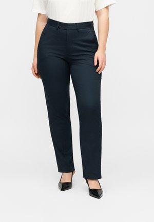 Kvinde iført sorte tætsiddende bukser med lige ben, hvid kortærmet top og sorte spidse hæle står mod en ensfarvet baggrund.