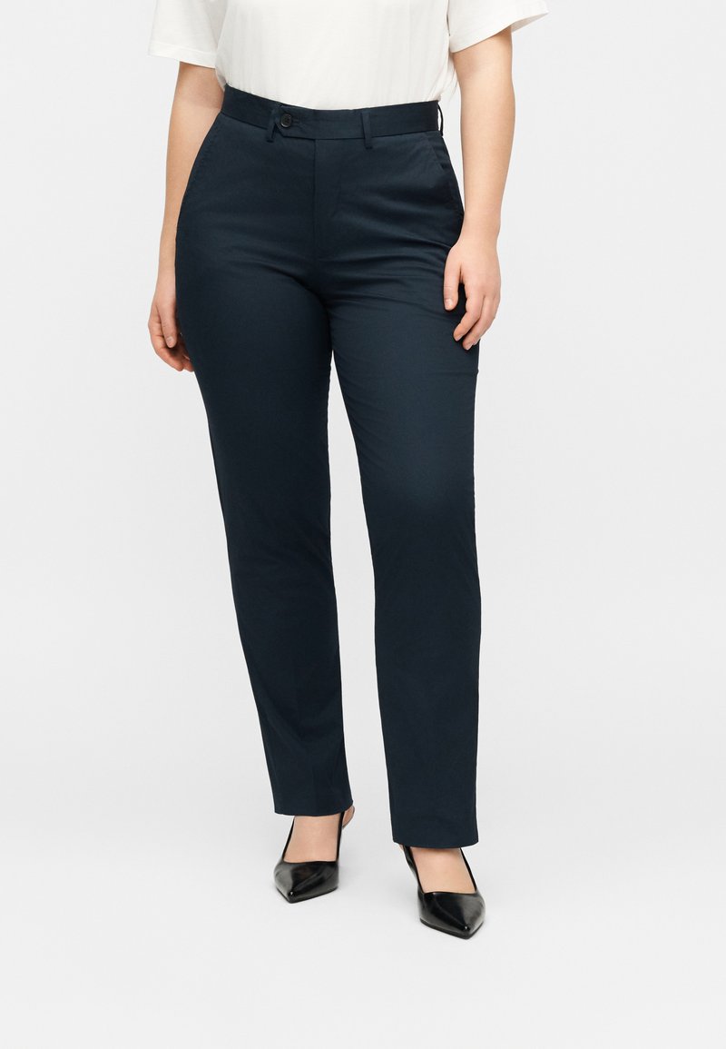 Γυναίκα που φοράει μαύρο εφαρμοστό παντελόνι με ίσια γραμμή, λευκό κοντομάνικο τοπ και μαύρες μυτερές γόβες, στέκεται μπροστά σε απλό φόντο.