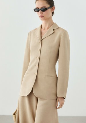 Femme portant un blazer beige sur-mesure et une jupe assortie, tenant un sac tressé, avec les cheveux lissés en arrière et des lunettes de soleil ovales foncées.