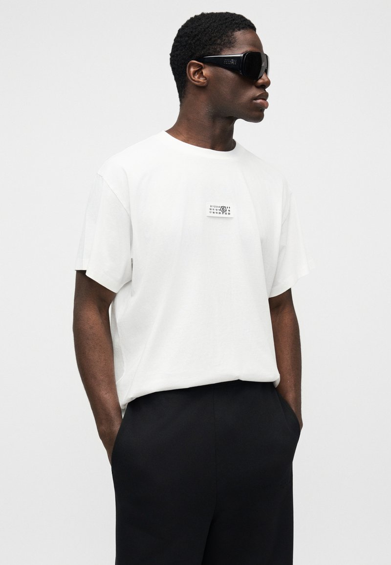 Hvid bomuld T-shirt med et lille grafisk motiv på brystet, kombineret med sorte bukser og store, sorte solbriller. Glat tekstur, afslappet pasform.