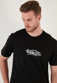 Melnas kokvilnas t-kreklu ar īsām piedurknēm, uz kura krūtīm ir balti grafiti stila burti "WORLDISER". Apaļa, klasiskā kakla izgriezuma forma.