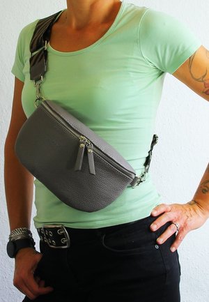 Sac bandoulière en cuir gris de forme arrondie, doté d'une fermeture zippée en haut et de deux pompons. Bandoulière réglable avec un motif zigzag.
