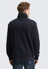 Marineblauwe pullover met hoge kraag, geribbelde textuur en lange mouwen. Afgewerkte manchetten en tailleband, gedragen met donkere spijkerbroek.