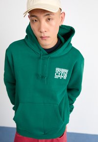 Green hooded sweatshirt with kangaroo pocket, white logo print "ADIDAS ORIGINALS SPORTS CLUB" on chest, textured fabric, drawstring detail.