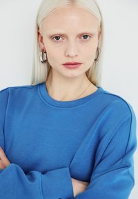 Kék pulóver, puha anyagból készült, kerek nyakkal és hosszú ujjal. A modellen ezüst fülbevalók vannak, és egyenes szőke haja.