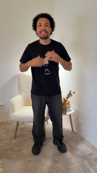 Junger Mann mit lockigem Haar, der lächelt, trägt ein schwarzes T-Shirt mit Grafik und Jeans, steht in einem Zimmer mit einem weißen Sessel und einem kleinen Tisch.