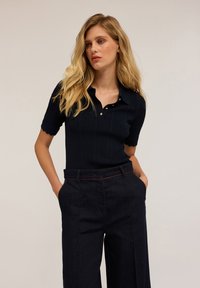 Polo shirt lavorato a maglia blu con maniche corte, caratterizzato da un motivo testurizzato e una chiusura con tre bottoni. Abbinato a pantaloni in denim scuro a vita alta.