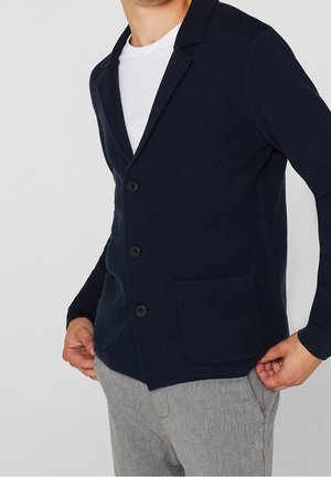 Marinblåstickad blazer med slag, fem knappar och en fuskficka. Bärs över en vit skjorta, ihop med ljusgråa mönstrade byxor.