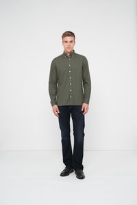 Camisa verde oliva abotonada, de manga larga, con detalle de bolsillo frontal; combinada con jeans azul oscuro y zapatos negros. El modelo está de pie sobre un fondo neutro.