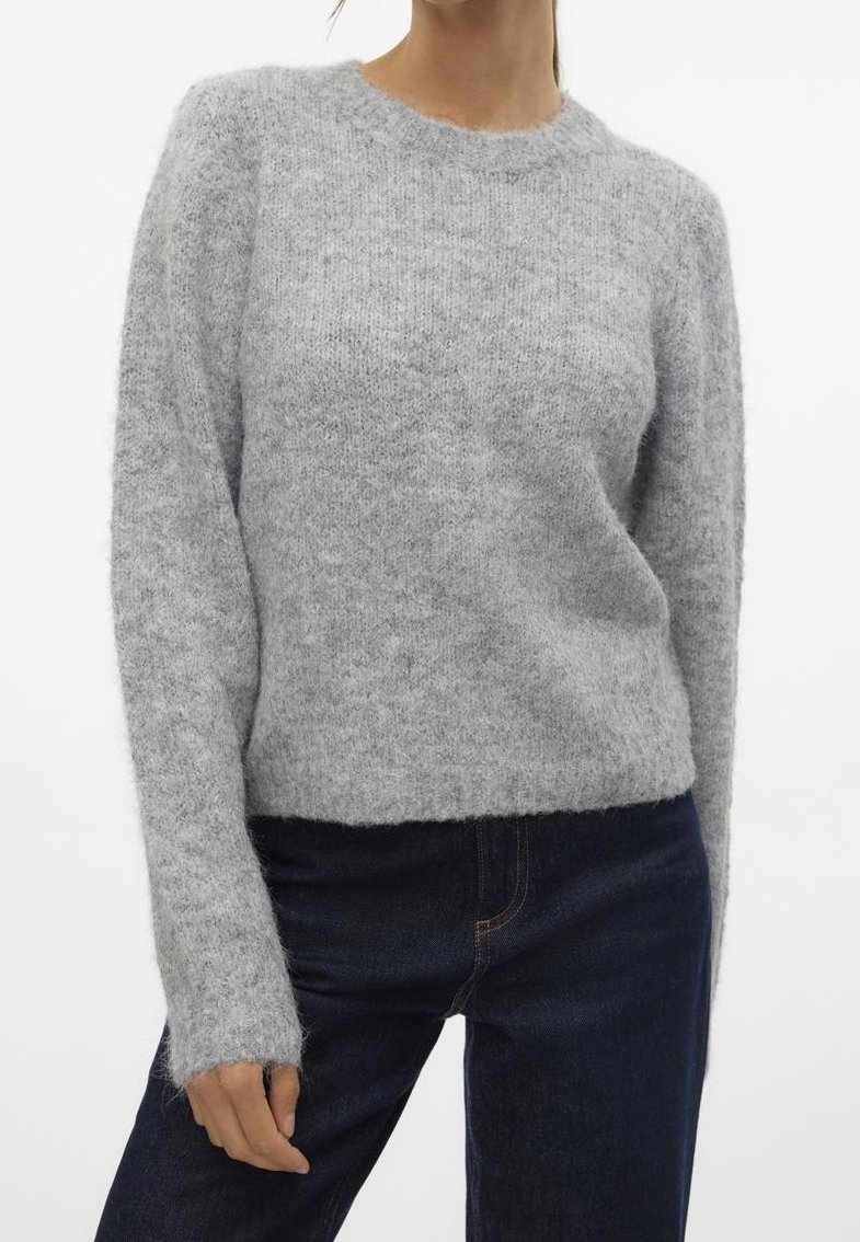 Femme portant un pull gris clair en maille pelucheuse à manches longues et un jean bleu foncé, debout devant un fond blanc uni.
