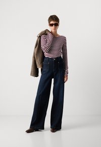 Bredde ben mørk denim jeans med gule sømmer, parret med en vinrød og hvit stripete langermet skjorte og en brun skinnjakke.