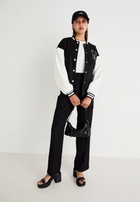 Veste varsity noir et blanc avec des poignets côtelés, haut blanc, pantalon noir à jambes larges, sandales à plateforme noires chunky, et un petit sac à main noir.