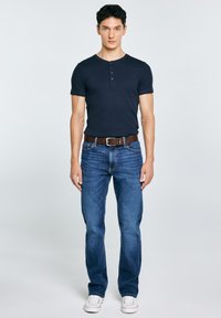 Ung mand står frontalt iført en marineblå kortærmet henley, blå jeans med brunt bælte og hvide sneakers foran en ensfarvet baggrund.