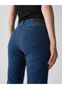 Jean en denim bleu foncé au design ajusté, avec des poches arrière et une ceinture noire. Le tissu a une texture lisse et une légère extensibilité.