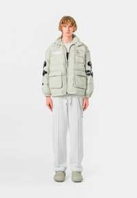 Chaqueta de plumas en verde claro con múltiples bolsillos frontales, detalles bordados en negro en las mangas, llevada sobre pantalones blancos y zapatos texturizados.