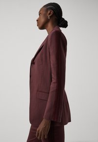Blazer ajustado en color burdeos con tejido texturizado. Presenta un cierre de un solo botón, bolsillo lateral y un detallado intrincado a lo largo de las mangas.