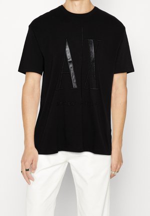 Mann trägt ein schwarzes Armani Exchange T-Shirt mit großem, geprägtem "AX"-Logo und weiße Hose, steht vor weißem Hintergrund.