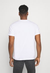 White cotton t-shirt with short sleeves, round neckline, and a small logo accent at the lower hem, paired with dark gray jeans.