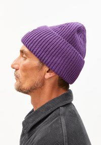 Lila gerippte Strickmütze mit abgerundetem Oberteil und enger Passform. Getragen zu einem dunklen Cordhemd, das Textur und Tiefe zeigt.