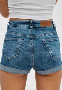 Pantalones cortos de mezclilla azul con cintura alta, dobladillos enrollados y un parche de cuero con marca en la parte trasera. Cuenta con dos bolsillos traseros y una textura desgastada.