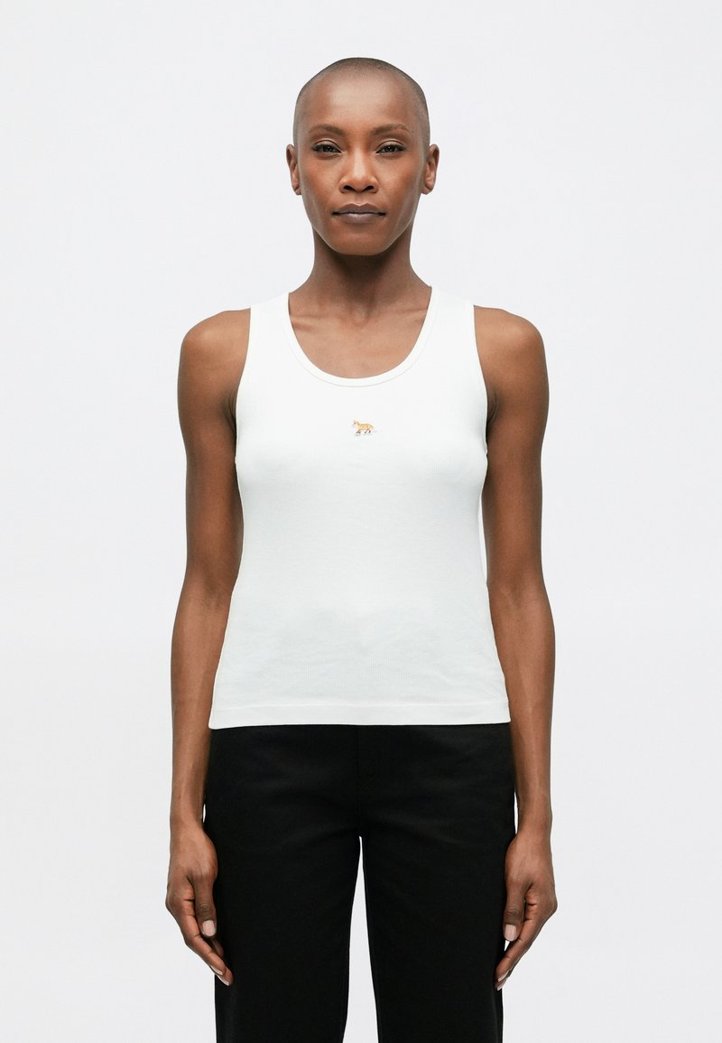 Balta marškinėlių su petnešomis, pagaminta iš tekstūrinio medvilnės, su mažuoju auksiniu gyvūnų logotipu centre. Modelis dėvi juodus kelnes.