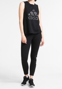 Czarny bezrękawnik sportowy z błyszczącym logo Adidas, w zestawie z czarnymi legginsami z białą grafiką. Model nosi czarne trampki.