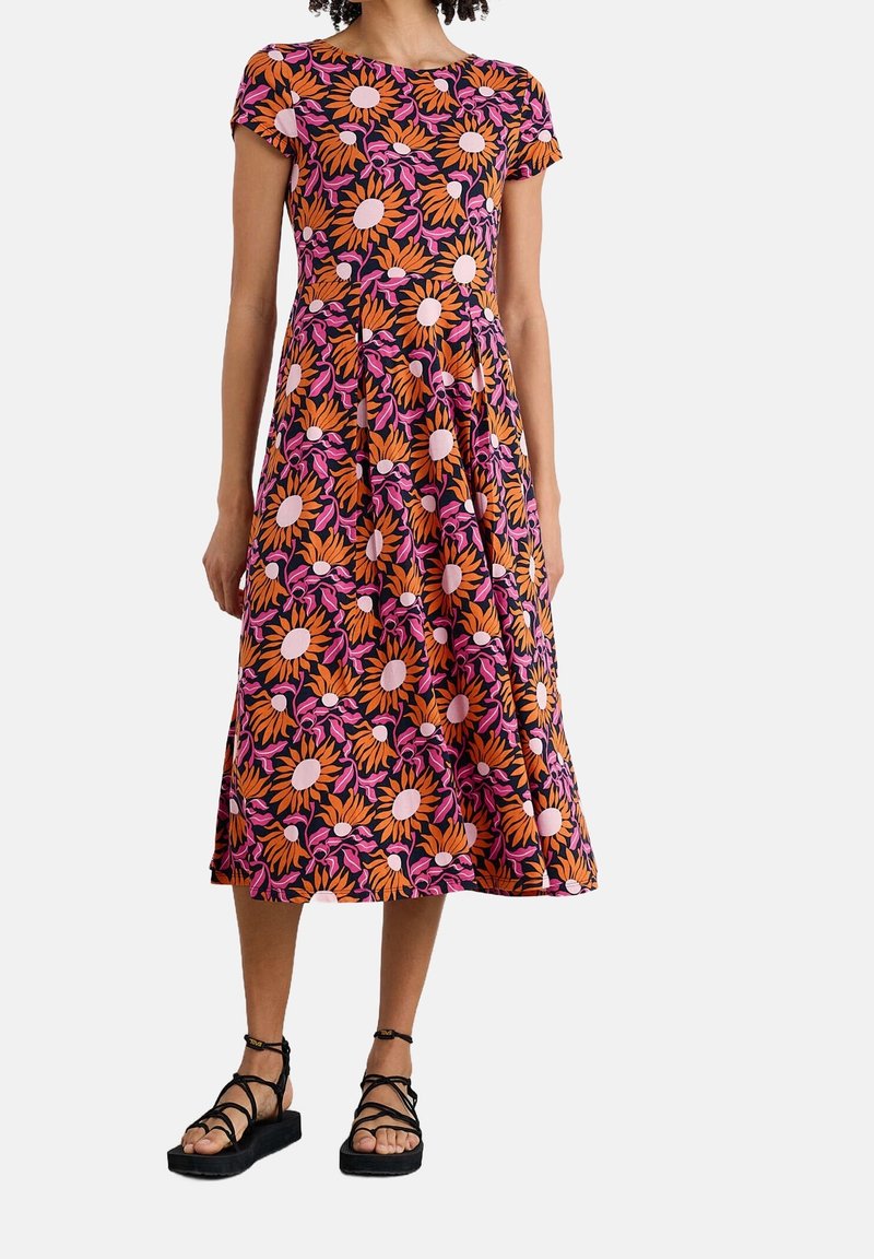 Femme portant une robe mi-longue avec un motif floral orange, rose et noir et des sandales plates noires à fines brides, debout devant un fond blanc.