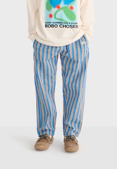 Une personne porte un pantalon à rayures verticales bleu et beige avec des chaussures mocassins beige et un haut à manches longues crème orné d'une impression colorée et de texte.