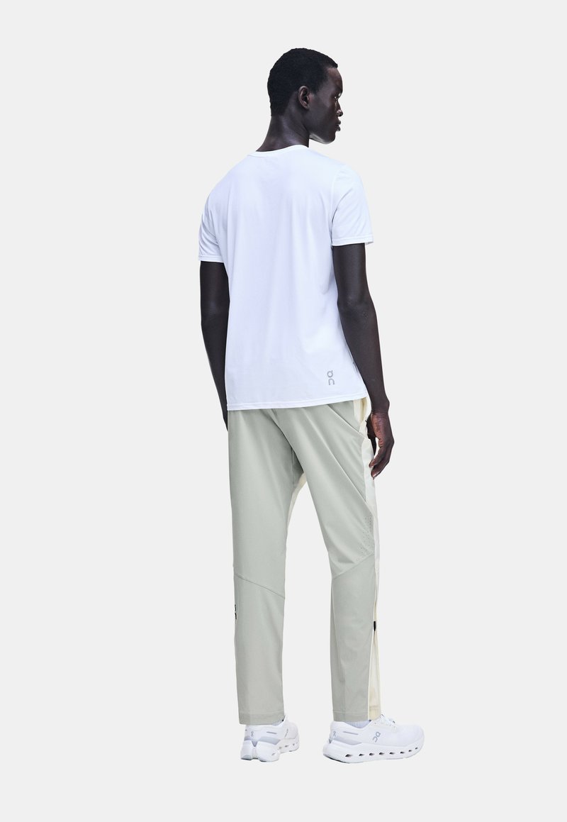 Homme portant un t-shirt blanc à manches courtes, un pantalon gris clair avec des panneaux latéraux crème, et des baskets blanches, debout de dos, la tête tournée vers la droite.
