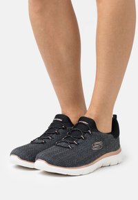 Baskets en noir et blanc avec un motif en chevron et des accents roses, dotées d'un matériau flexible, de lacets et d'une semelle blanche.