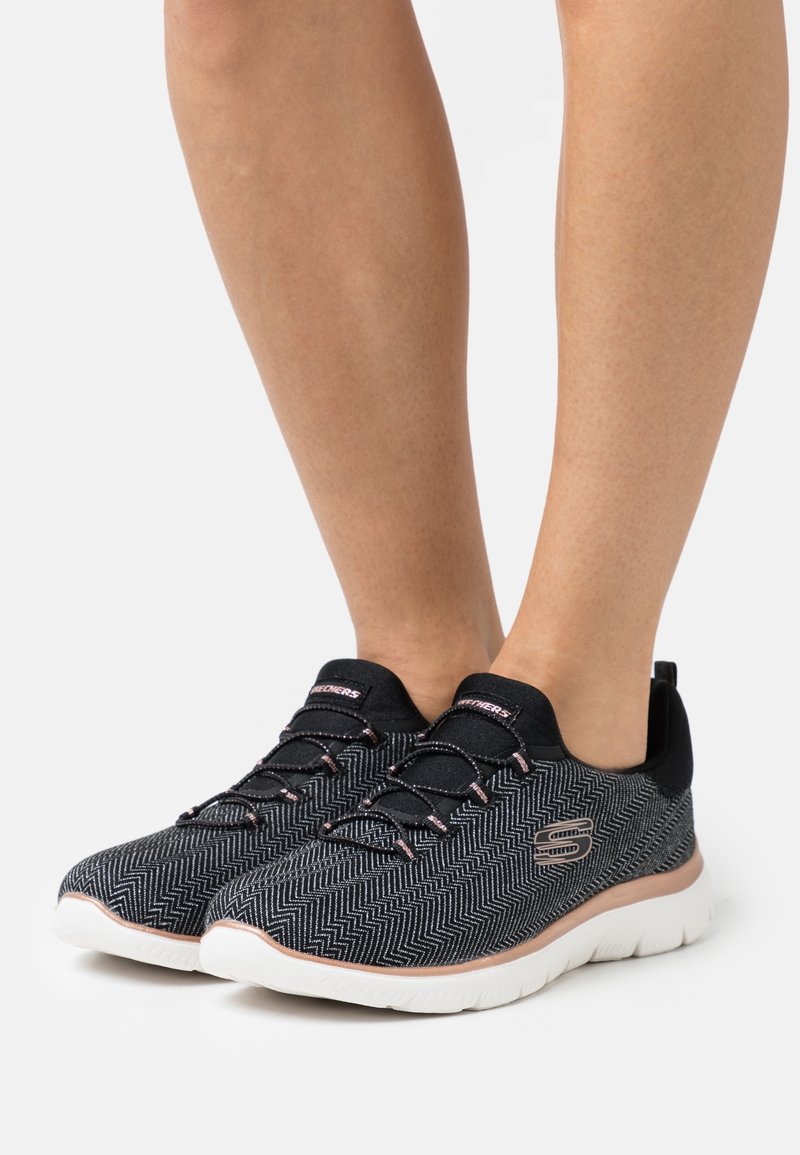 Baskets en noir et blanc avec un motif en chevron et des accents roses, dotées d'un matériau flexible, de lacets et d'une semelle blanche.